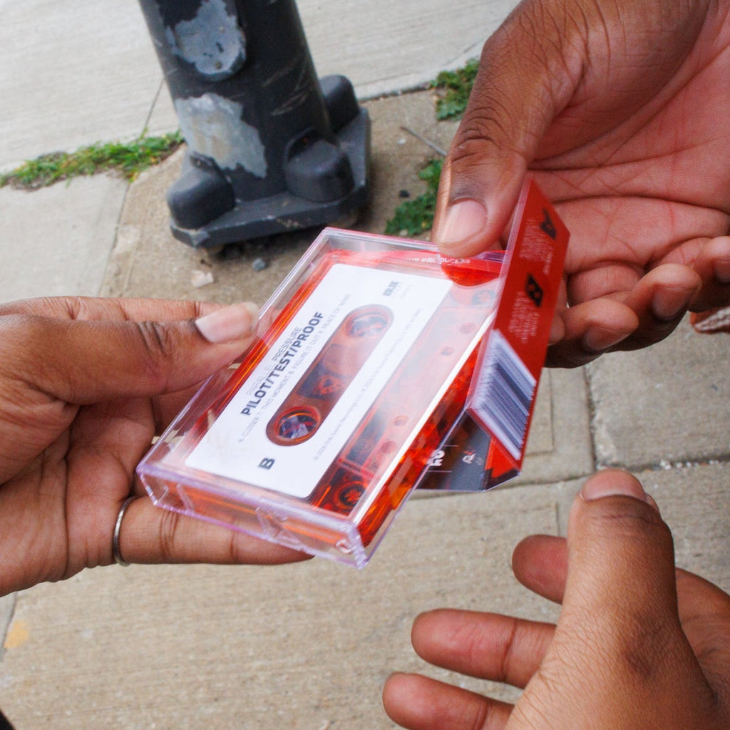 A person holding a clear plastic cassette case with a red-and-white printed insert, shown in an outdoor street setting with hands and a sidewalk background.
