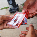 A person holding a clear plastic cassette case with a red-and-white printed insert, shown in an outdoor street setting with hands and a sidewalk background.